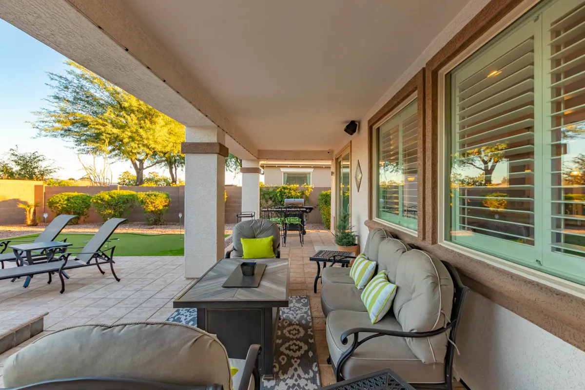 A cozy patio area with comfortable seating, plush decorative pillows, a dining table, and lush greenery all around. The space is set up for relaxation or al fresco dining, with touches that invite guests to linger, such as the fire pit and the view of the surrounding landscaping.