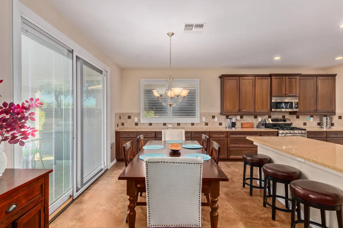 A stylish dining area adjacent to a kitchen island, featuring a welcoming dining table, a modern chandelier overhead, and sliding glass doors that allow sunlight to pour in. The space is designed for casual meals and lively gatherings.