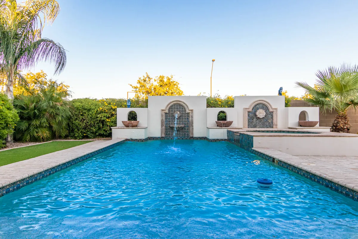 A serene backyard scene featuring a sparkling swimming pool, decorative wall fountain, lush greenery, and tall palm trees. This space is ideal for swimming, relaxing, or socializing outdoors under the Arizona sun.