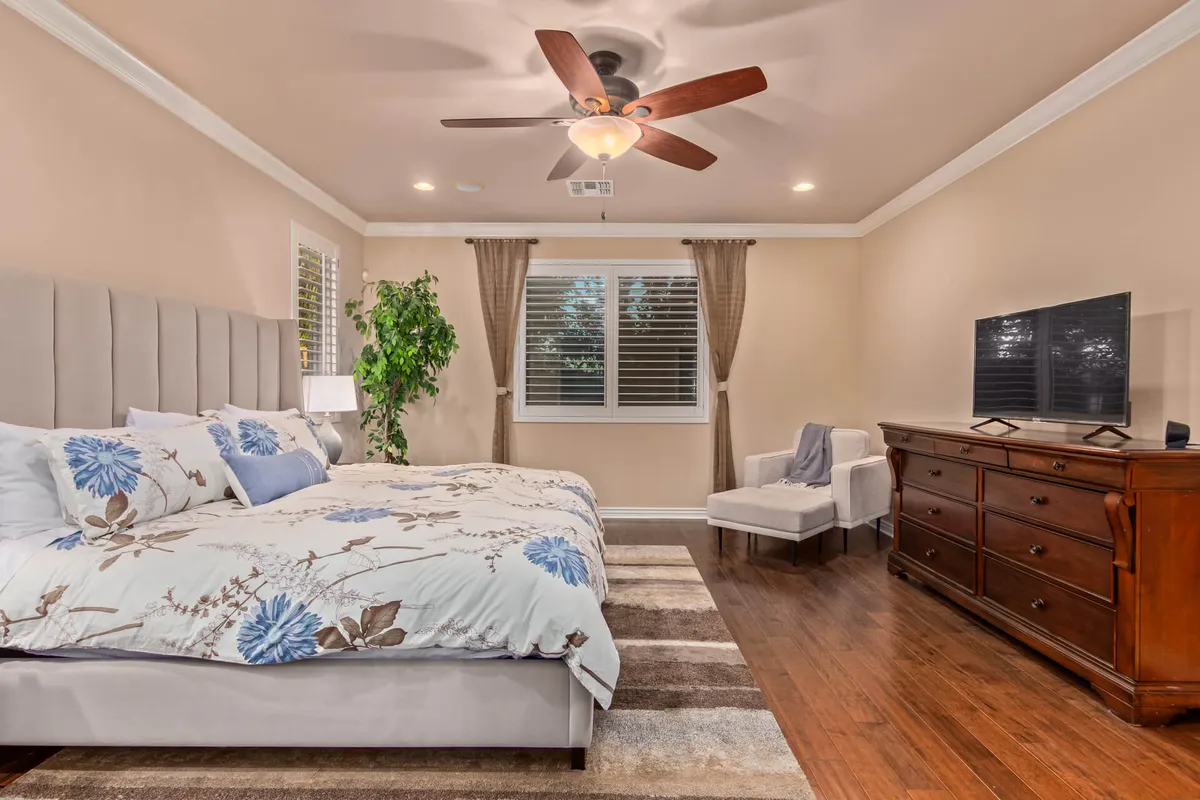 A comfortable bedroom featuring a large bed with floral bedding, a wooden dresser, a cozy armchair, and a potted plant. Natural light filters in, creating a serene and restful ambiance that speaks to both comfort and style.