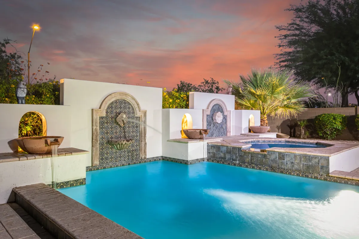 A serene exterior view of a swimming pool surrounded by lush greenery and palm trees, with sunset hues in the sky. Lounge chairs and water features create a relaxing, luxurious retreat after a day of exploration.