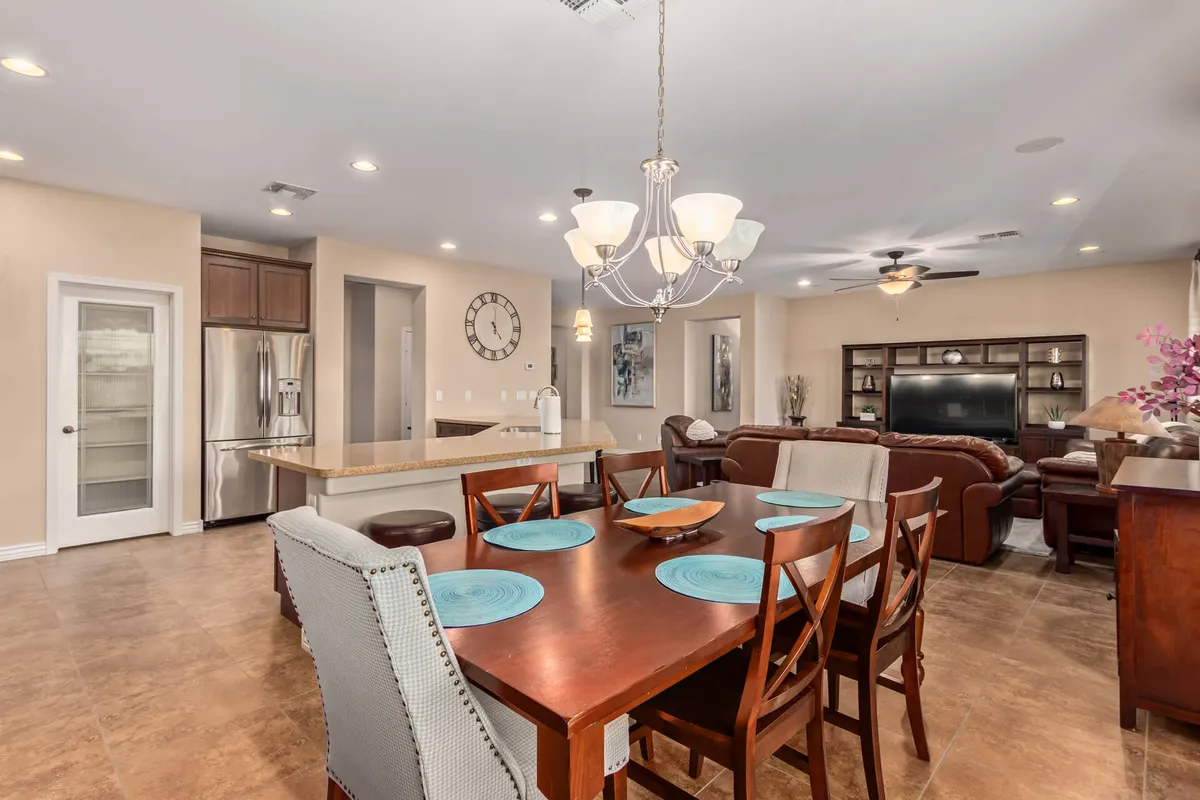 An open-plan setting showing a dining table with blue plates set for dinner, a sparkling chandelier, and a glimpse of the living room seating area. The large clock and thoughtful decor enhance the sense of warmth and hospitality.