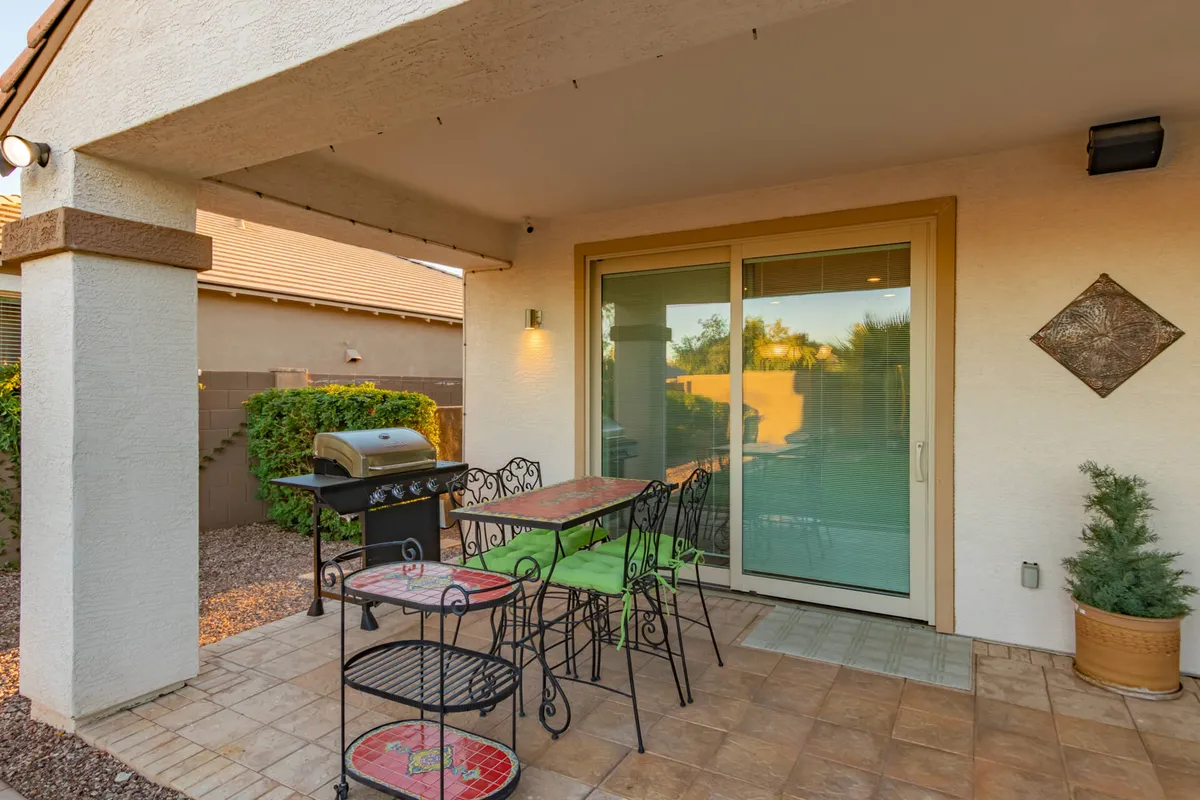 An inviting backyard patio featuring an outdoor dining set and a grill adjacent to a sliding glass door. The area is set up for al fresco meals, surrounded by greenery, and bathed in warm, late-day light ideal for gatherings.