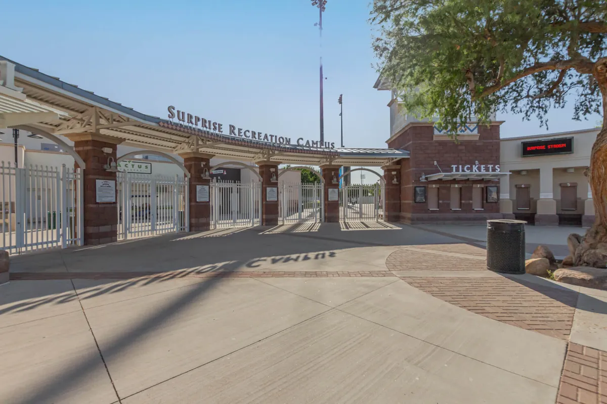 The entrance gates and ticket booth to a sports venue, framed by surrounding trees. The scene is lively and welcoming, suggesting easy access and the excitement of attending major sporting events with friends and family.