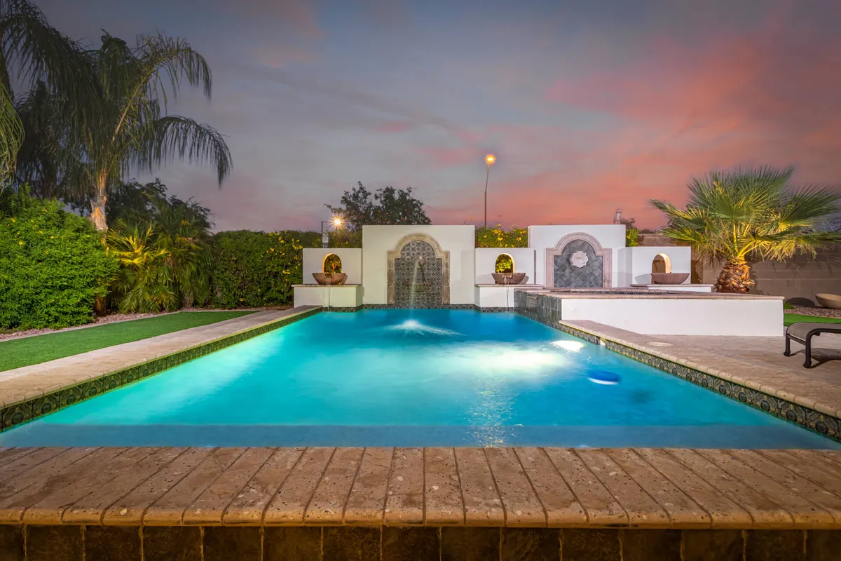 A serene backyard scene featuring a sparkling swimming pool, decorative fountain, lush greenery, and towering palm trees. The landscaping is distinctly Southwestern, with rocks and desert-adapted plants framing the water, and the area is bathed in golden sunset light, evoking a peaceful and inviting atmosphere.