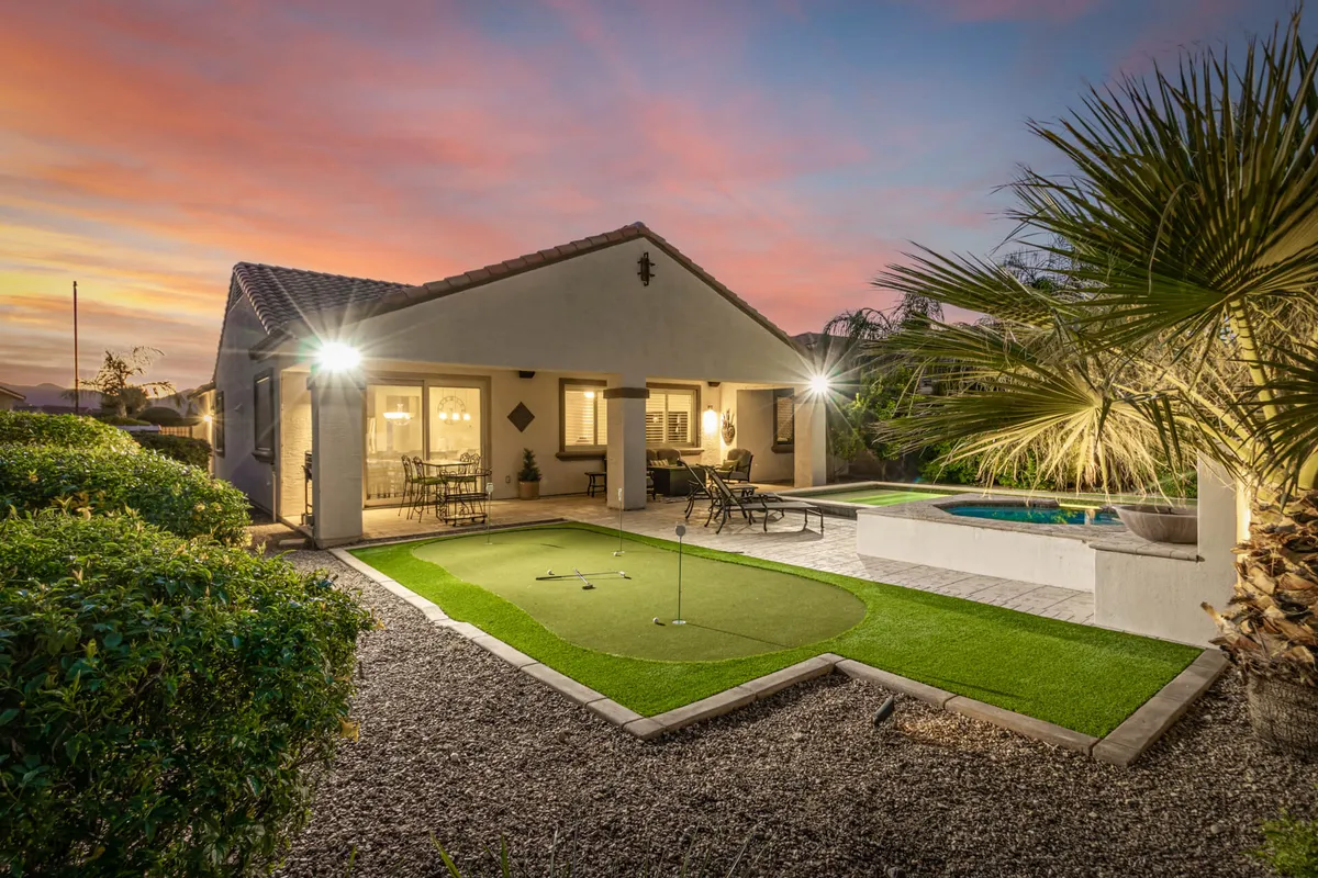 A beautifully landscaped backyard featuring a golf putting green, sparkling swimming pool, outdoor seating area, and tropical palm trees. The setting is bathed in warm sunlight and offers views ideal for both golf practice and leisurely relaxation.