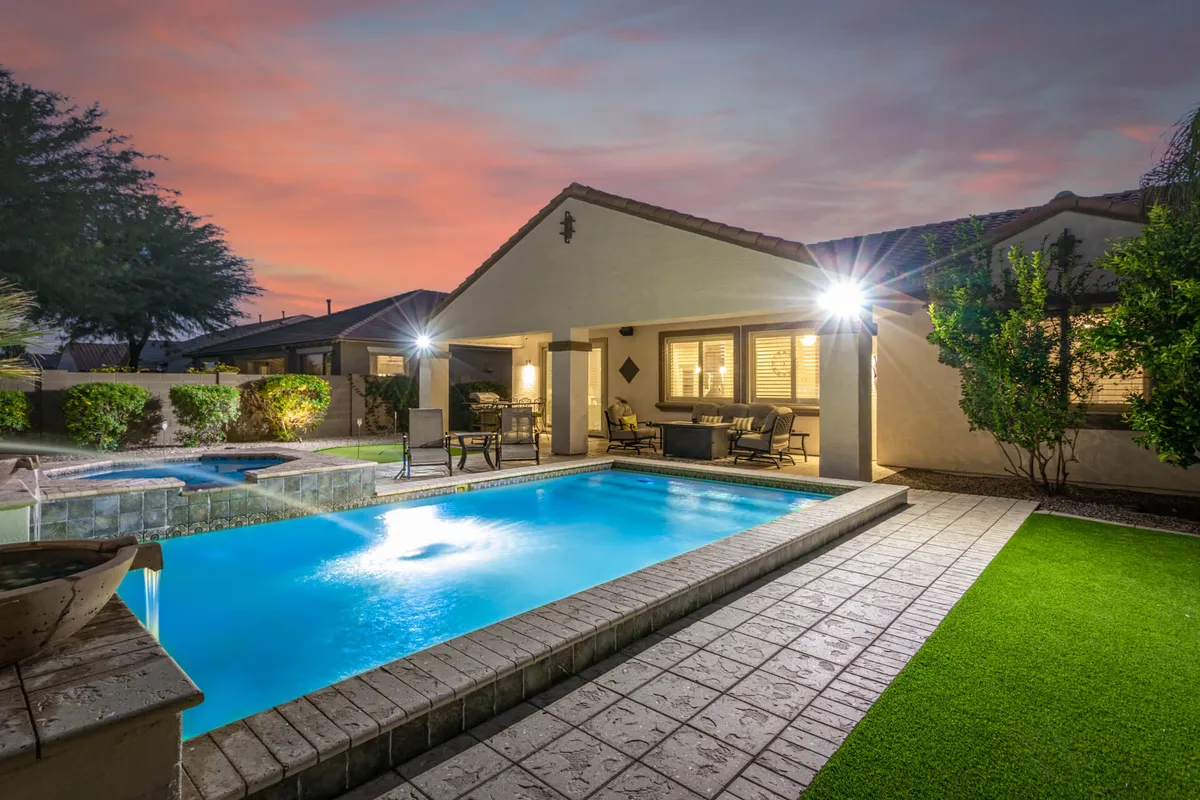 The image shows a sun-drenched backyard oasis with a sparkling swimming pool, an inviting jacuzzi, comfortable outdoor seating surrounded by lush palm trees, and stylish Southwestern landscaping. The setting is perfect for swimming, relaxing, or gathering with friends and family under the Arizona sky.