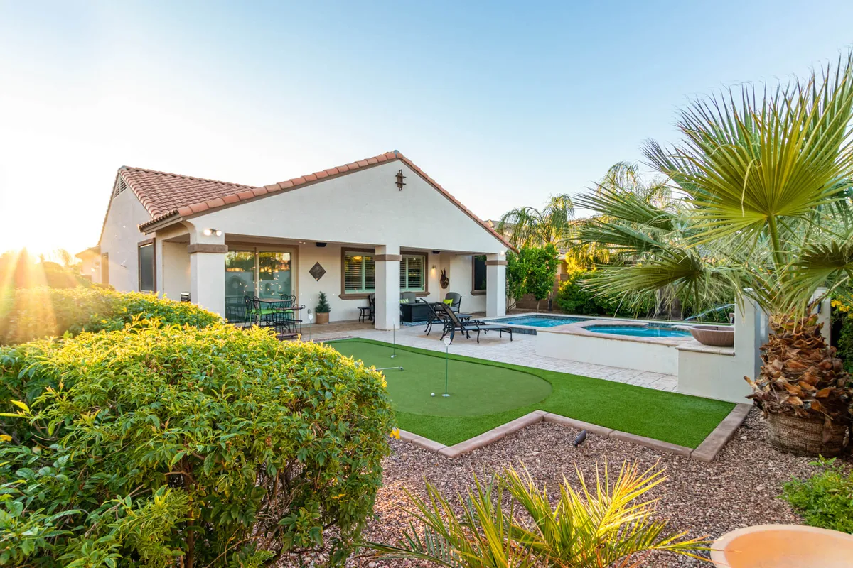 A vibrant backyard view showcasing a sparkling swimming pool, a shaded patio seating area, a putting green, and lush, vibrant greenery. This space is clearly designed for both relaxation and entertainment, with options for sunbathing, swimming, playing, or gathering with friends and family at sunset.